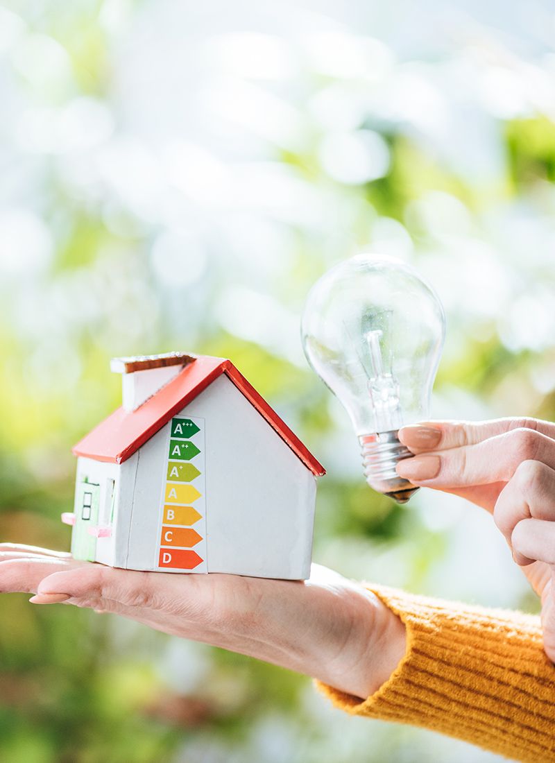 Primer plano de una mano que sujeta una casa con las diferentes certificaciones energéticas y en la otra mano sujeta una bombilla.